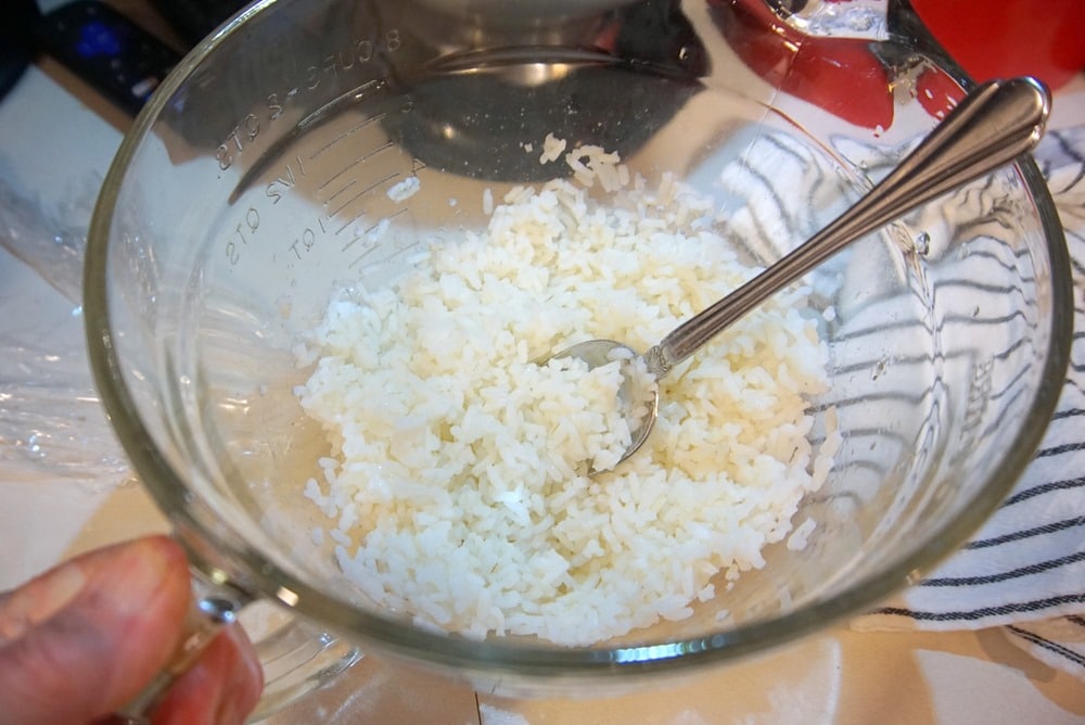 A glass mixing bowl with cooked white rice and a metal spoon inside. The bowl is held by a hand and sits on a striped cloth, ready for mixing into a classic Hopping John dish, with kitchen items blurred in the background.