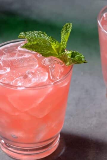 A glass filled with a pink iced drink, garnished with a fresh mint sprig, sits on a gray surface. Another Sweetheart Cocktails creation is partially visible in the background.