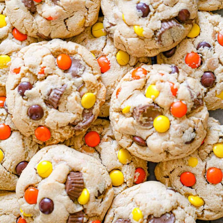 A close-up of several large baked desserts filled with pieces of peanut butter cups and colorful orange, yellow, and brown candy-coated chocolates. The cookies have a soft, chunky texture.