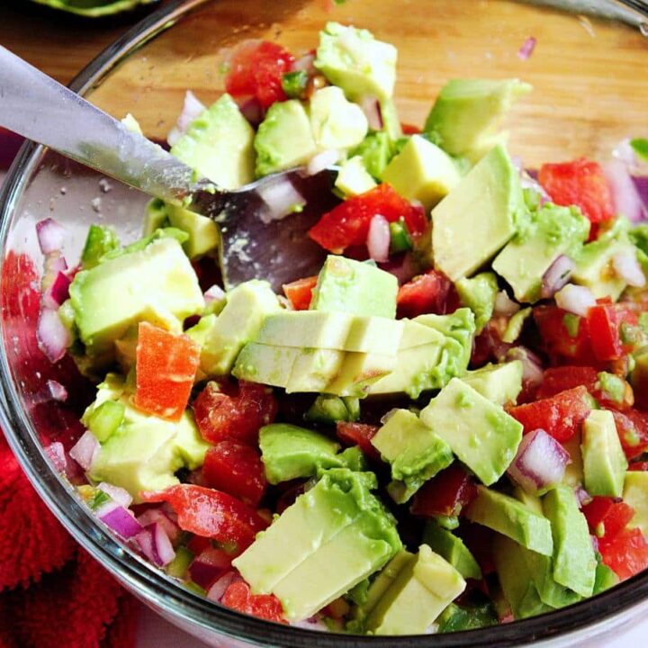 A glass bowl filled with chunky jalapeno guacamole, made with avocado, diced tomatoes, and chopped red onions. A metal spoon is in the bowl, and a red cloth is nearby on the counter.
