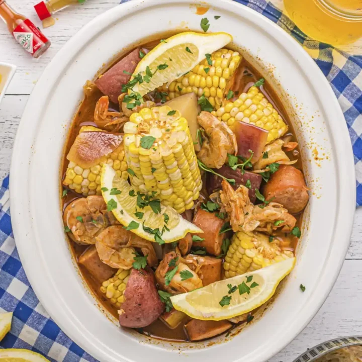 Oval white dish filled with a seafood boil featuring crab and shrimp recipes, corn on the cob, potatoes, sausage, and clams, garnished with lemon wedges and parsley. Surrounded by bread, lemon slices, drinks, and hot sauce bottles on a checkered cloth.