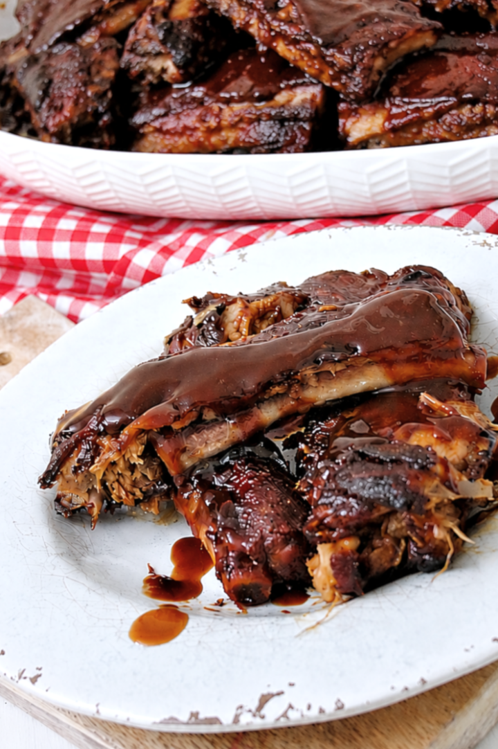 A plate with several pieces of Instant Pot Dump and Go Country Style Ribs covered in dark, glossy sauce sits on a white surface, with a red and white checkered cloth and a serving tray of more ribs in the background.