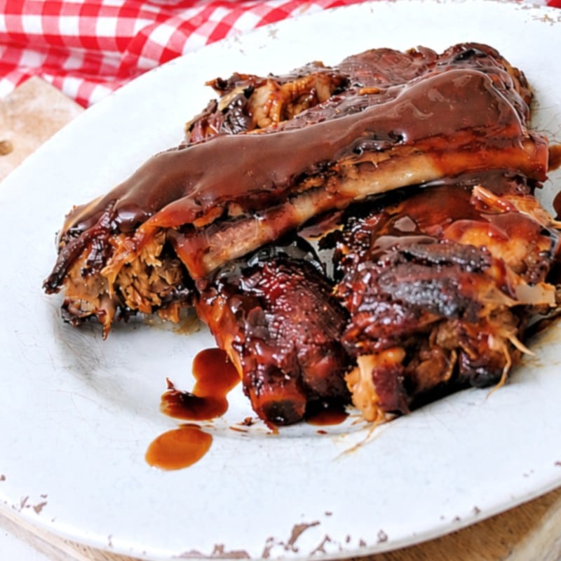 Instant Pot Dump and Go Country Style Ribs coated in a glossy, dark sauce are served on a white plate, with a red and white checkered cloth in the background.