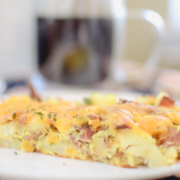 A close-up of a cheesy ham and potato breakfast casserole, perfect for egg recipes lovers, on a white plate, with a blurred mug of coffee in the background.