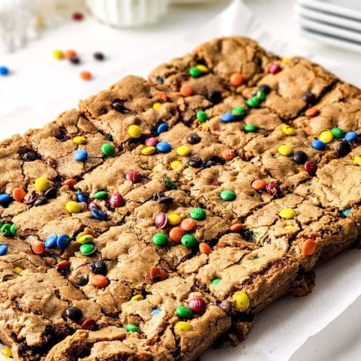 A large rectangular M&M’s Cookie Bar topped with colorful candy-coated chocolates, cut into squares and placed on parchment paper, with scattered candies around on a white surface.