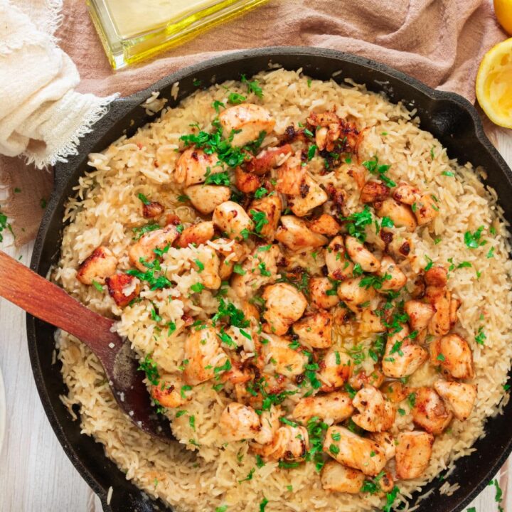 A skillet filled with creamy lemon chicken and rice, topped with seasoned, browned pieces and garnished with chopped parsley. A wooden spoon rests in the pan, surrounded by a towel, fresh lemon, and a bottle of oil on a light surface.