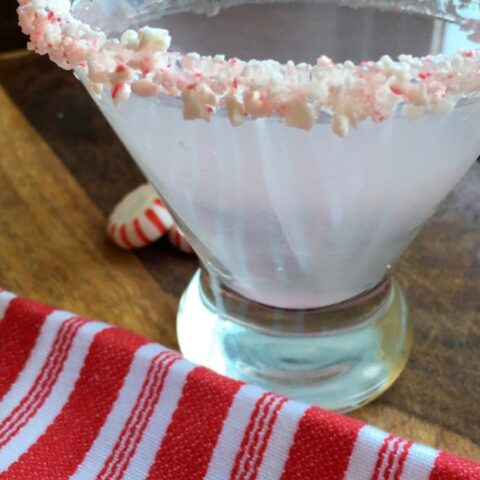 A martini glass with a crushed peppermint rim, filled with a pale pink cocktail, sits on a wooden surface next to a red and white striped cloth and a peppermint candy—perfect for serving festive cocktails.