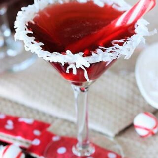 A festive cocktail in a martini glass with a shredded coconut rim, garnished with a small red and white candy cane. The glass sits on a table with a red polka dot ribbon and peppermint candies nearby.