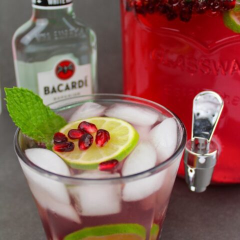 A glass of iced festive cocktail with lime slices, pomegranate seeds, and a mint sprig sits in front of a Bacardi white rum bottle and a large dispenser of pink punch garnished with lime and pomegranate.