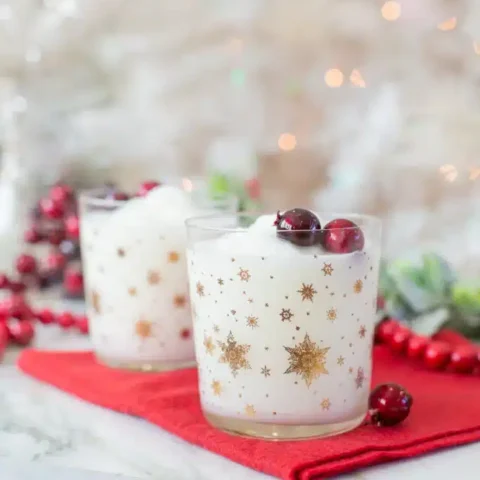 Two festive cocktails in gold snowflake glasses are filled with a white frothy drink and topped with red cranberries. They rest on a red napkin, surrounded by blurred holiday decor in the background.