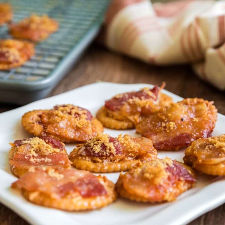 A white plate holds brown sugar bacon crackers, their sweet and savory topping glistening. In the background, more of these irresistible crackers rest on a cooling rack beside a striped cloth.