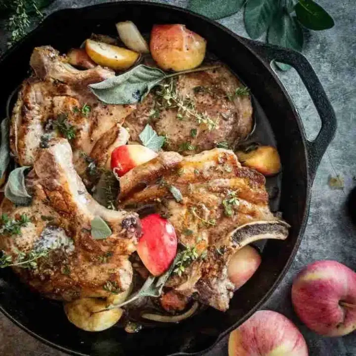 A cast iron skillet filled with cooked pork chops, garnished with herbs and surrounded by slices of apples. This delicious dish draws inspiration from classic dishes with honey. Fresh apples and herbs are also visible on the gray surface nearby.