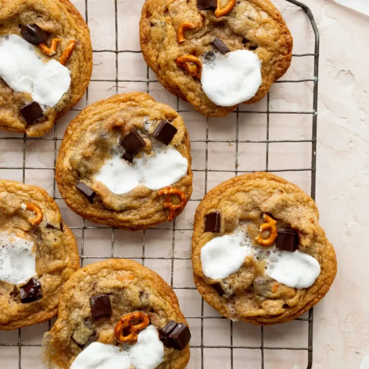 Cookies with melted marshmallows, chocolate chunks, and pretzel pieces are cooling on a wire rack atop a light surface—perfect handheld treats for a crowd. Some cookies rest on parchment paper, ready to share.
