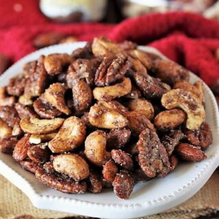 A white plate filled with assorted roasted nuts covered in a savory glaze, showcasing one of the finest nut dishes, sits on a wooden table with a red cloth and a can blurred in the background.