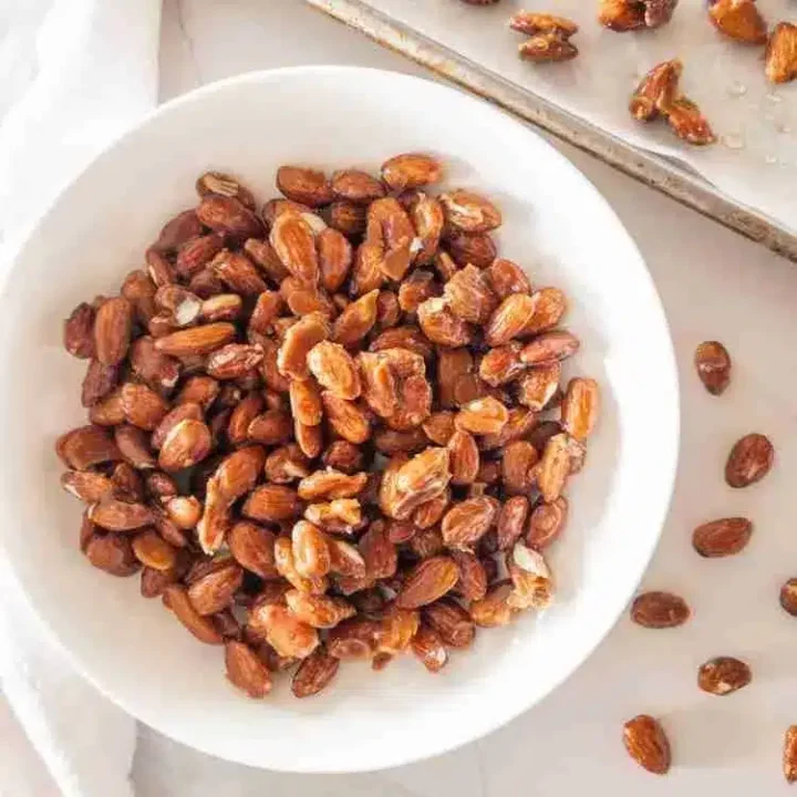 A white bowl filled with roasted, shelled peanuts—a classic choice for nut dishes—sits on a white surface. Some peanuts are scattered around the bowl and on a metal tray in the background.