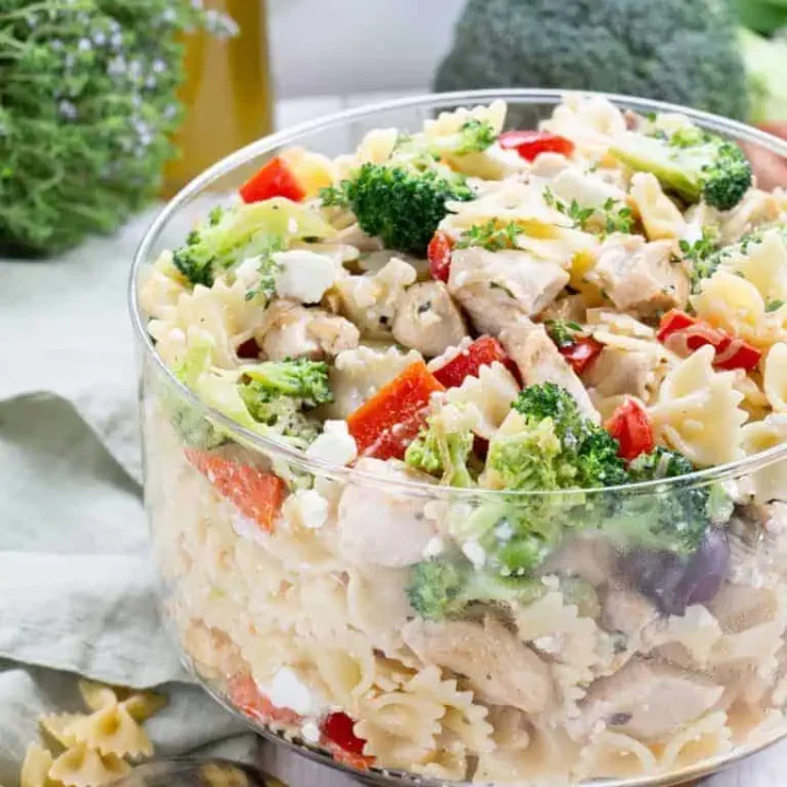 A glass bowl filled with pasta salad—bowtie pasta, broccoli, red bell peppers, chicken pieces, and crumbled cheese—perfect for Potluck Salads, set on a light surface with vegetables and olive oil in the background.