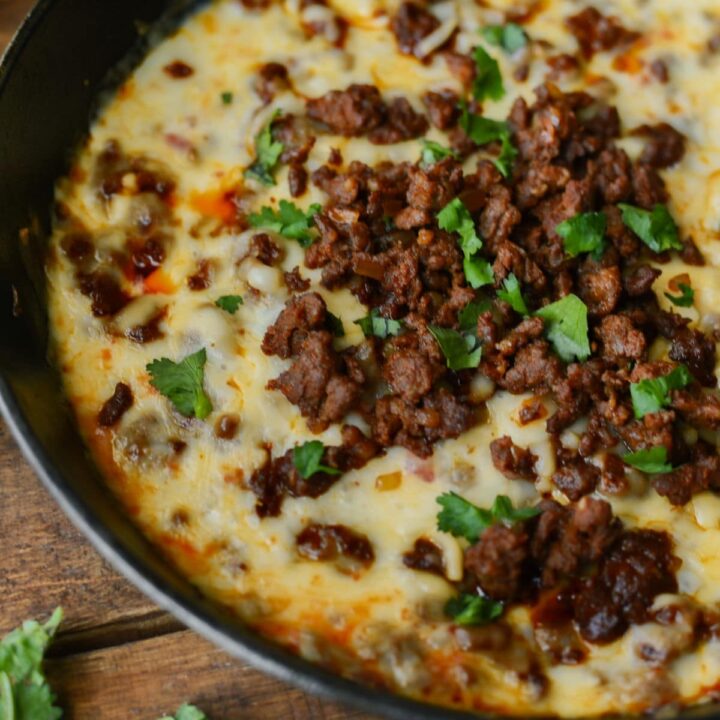 A skillet filled with queso fundido, topped with cooked ground beef and garnished with fresh cilantro, placed on a wooden surface.