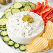 A bowl of creamy Dill Pickle Dip topped with pickle slices and dill, surrounded by sliced cucumbers, red bell pepper strips, and ridged potato chips on a white plate.