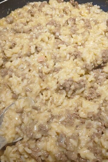 A close-up of a skillet filled with a creamy mixture of ground beef, rice, and melted cheese, with a metal spoon partially visible on the left side.