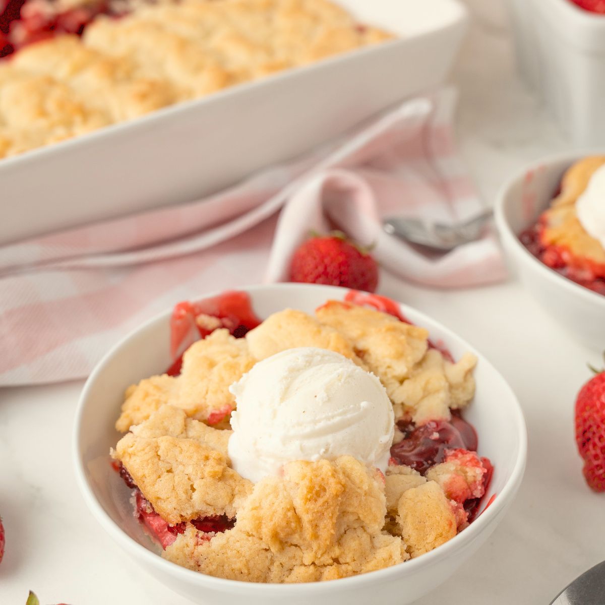 A bowl of strawberry dump cake cobbler topped with vanilla ice cream sits on a table next to fresh strawberries, with a baking dish and a pink-striped napkin in the background.