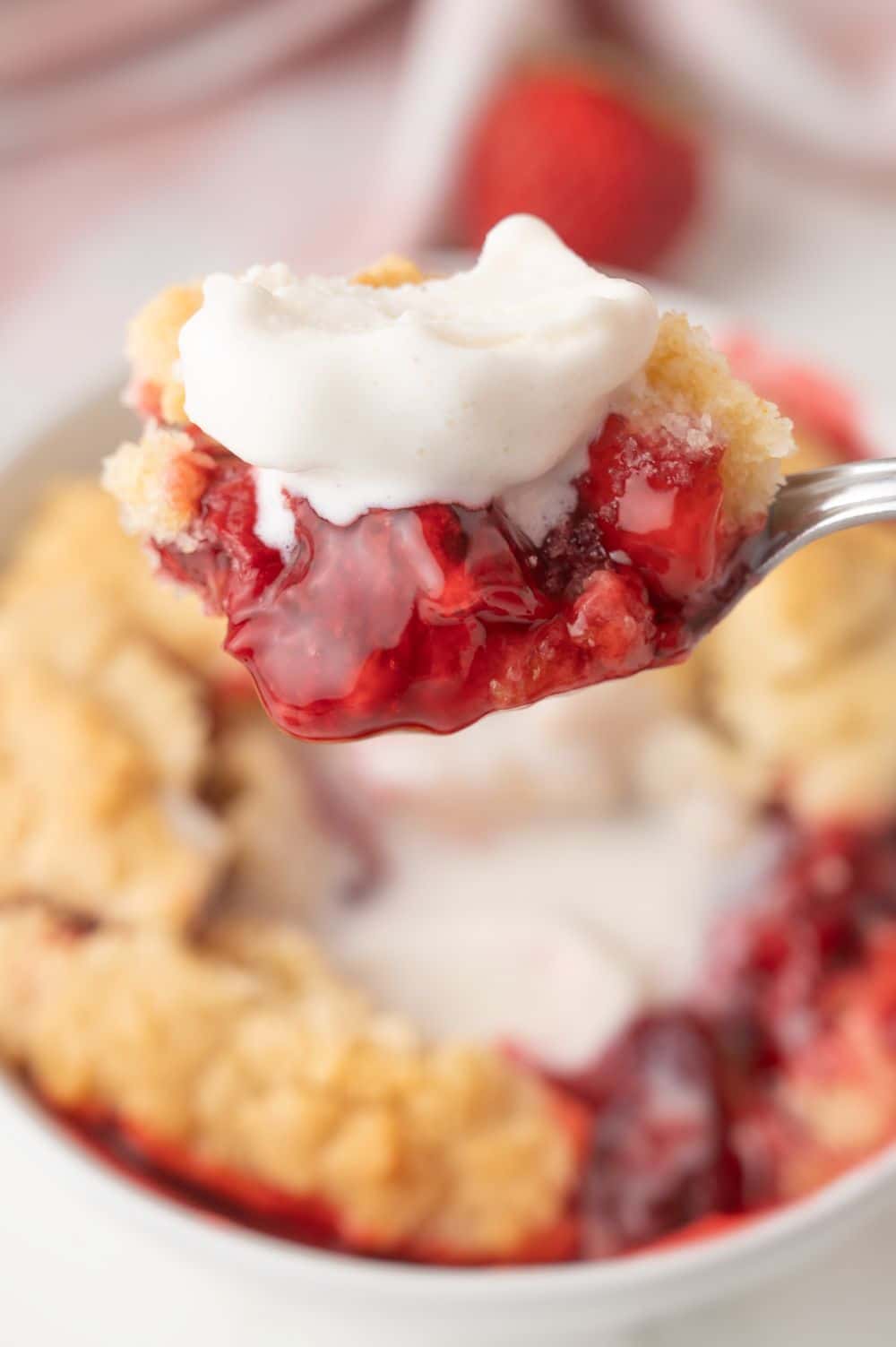 A close-up of a spoonful of berry cobbler, similar to a strawberry dump cake, topped with whipped cream, held above a bowl filled with more cobbler and juicy berry filling. A strawberry is blurred in the background.