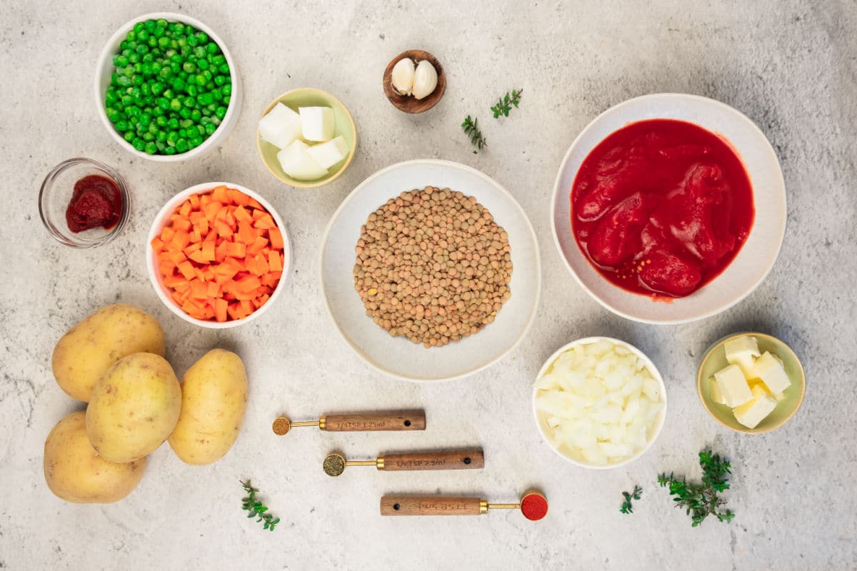 Overhead view of ingredients for Lentil Stew on a light surface: peas, diced carrots, tomato paste, potatoes, lentils, whole tomatoes in sauce, chopped onions, butter cubes, garlic cloves, salt, and fresh thyme.