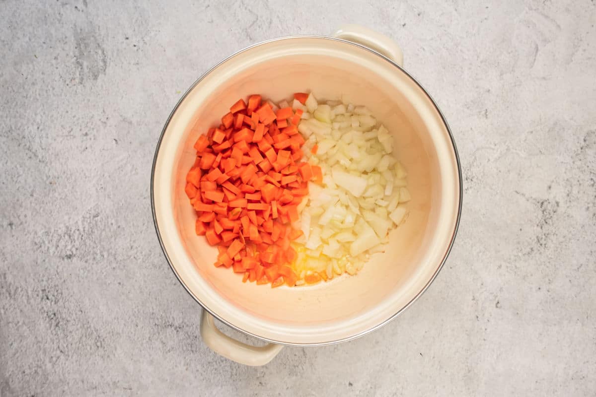 A large cream-colored pot with diced carrots and onions side by side on the bottom, ready to start a hearty Lentil Stew, viewed from above on a light gray surface.
