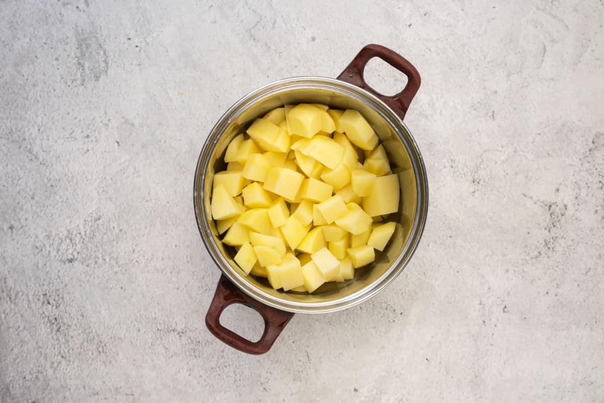 A metal pot with brown handles filled with peeled and evenly diced raw potatoes, ready to be added to a hearty lentil stew, sits on a light gray textured surface.