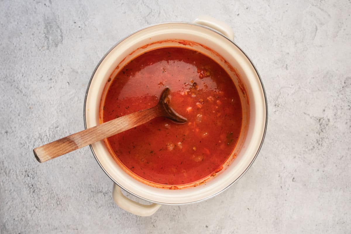 A white pot filled with a rich lentil stew, featuring tomato chunks and visible herbs, sits on a light grey surface. A wooden spoon rests inside the pot, partially submerged in the hearty mixture.