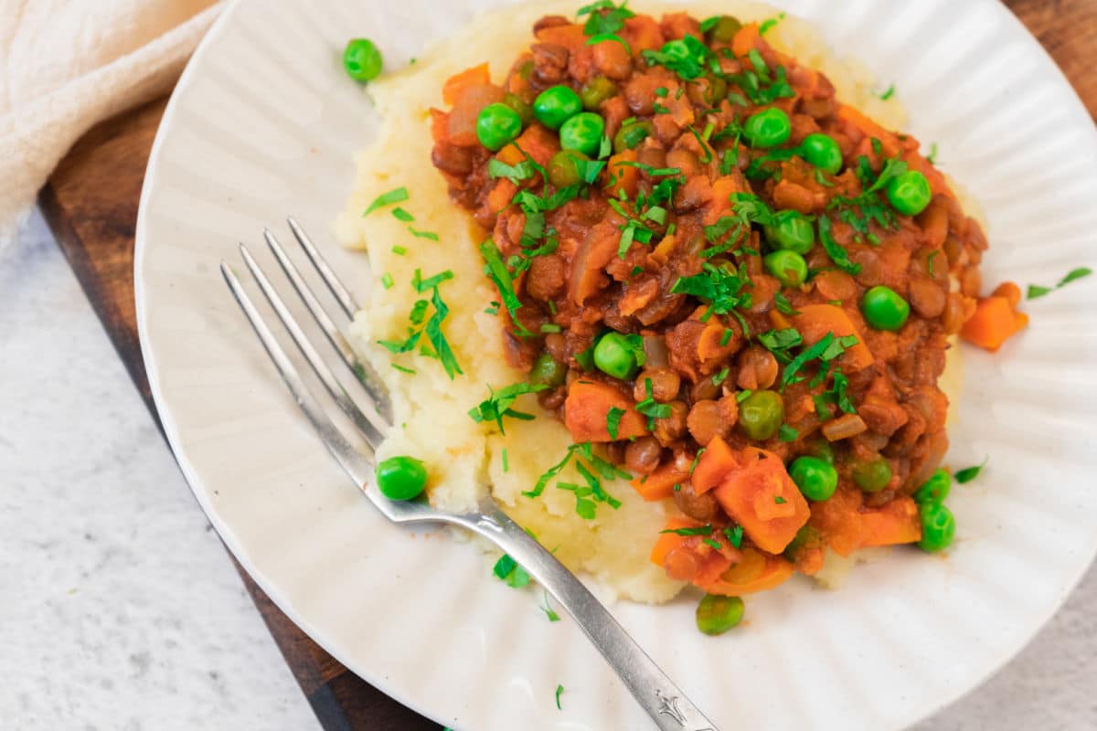 A plate of mashed potatoes topped with hearty Lentil Stew, filled with peas and carrots, and garnished with fresh herbs. A fork rests on the side of the plate.