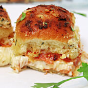 Close-up of a baked slider with a golden, crispy top, filled with melted cheese and slices of pepperoni, garnished with a sprig of fresh parsley on a white plate.