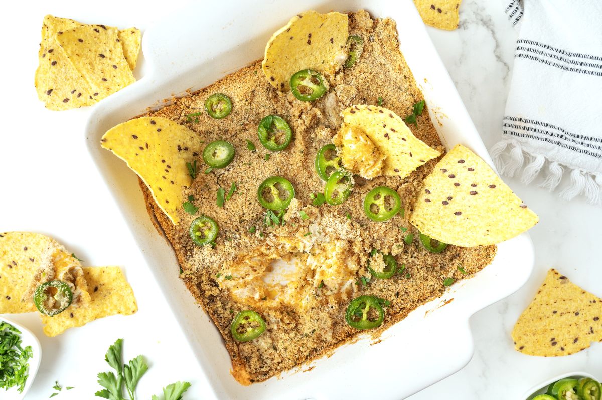 A square baking dish filled with creamy Jalapeno Popper Dip, topped with breadcrumbs and sliced jalapeños, is surrounded by tortilla chips on a white surface with a striped towel and fresh herbs nearby.