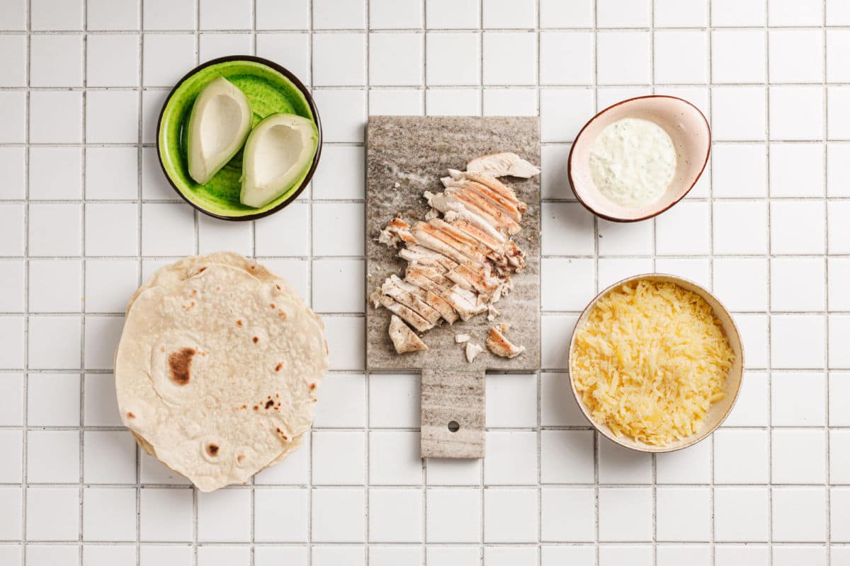Five ingredients for Chicken Avocado Ranch Burritos laid out on white tile: sliced grilled chicken on a cutting board, shredded lettuce, tortillas, avocado halves, a bowl of shredded cheese, and a small bowl of creamy ranch sauce.