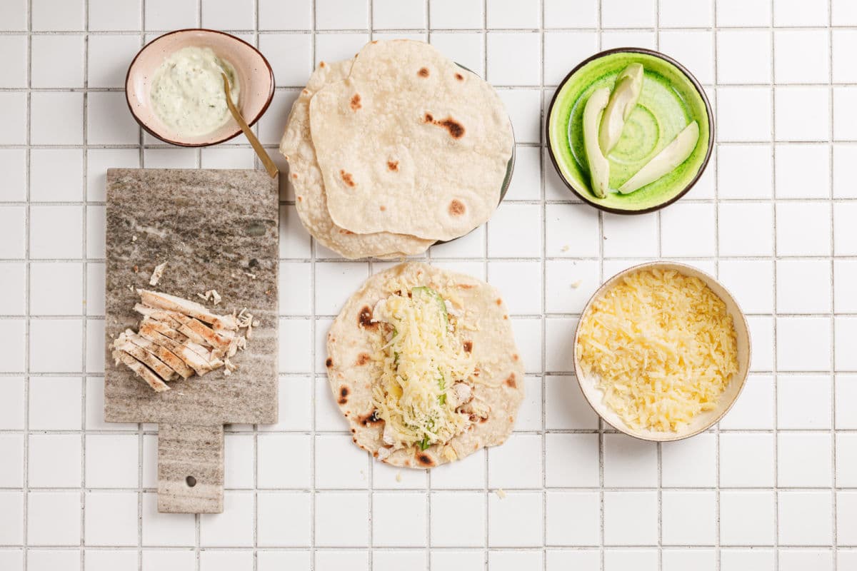 A flat lay of taco ingredients on a white tiled surface: grilled chicken strips on a small board, tortillas, shredded cheese, and sliced avocado in a bowl—perfect for assembling Chicken Avocado Ranch Burritos with a creamy sauce on the side.