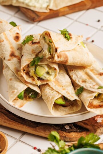 A plate of Chicken Avocado Ranch Burritos filled with vegetables and herbs, garnished with fresh cilantro, sits on a wooden board atop a tiled surface with scattered peppercorns and salt.