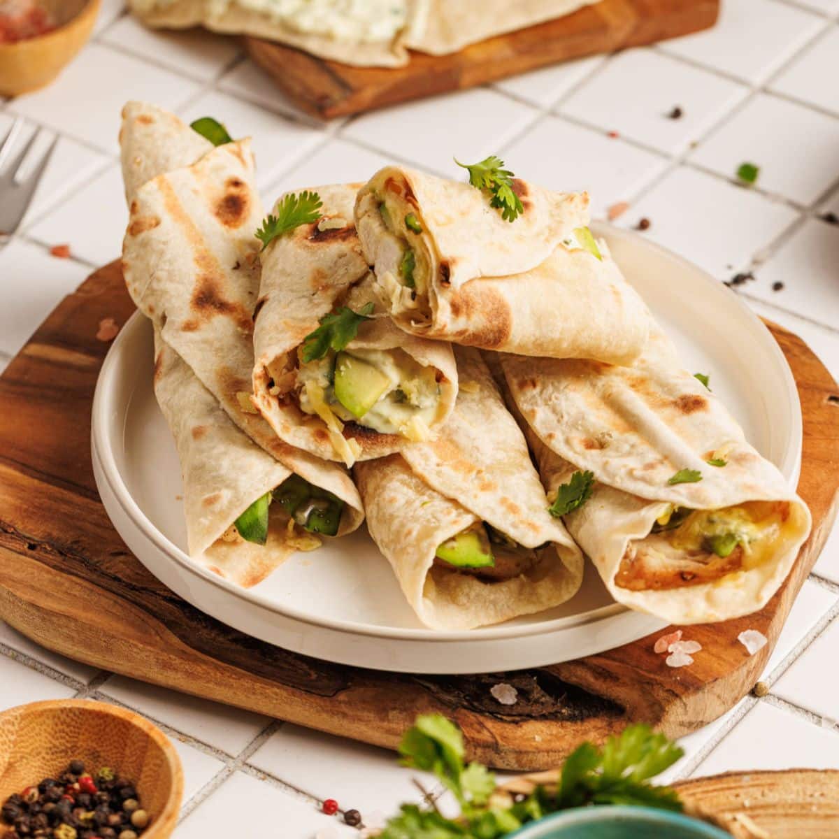 A plate of Chicken Avocado Ranch Burritos filled with vegetables and herbs, garnished with fresh cilantro, sits on a wooden board atop a tiled surface with scattered peppercorns and salt.