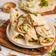 A plate of rolled flatbreads, inspired by Chicken Avocado Ranch Burritos, filled with vegetables and herbs sits on a wooden board. Other flatbreads, a bowl of sauce, grated cheese, and fresh cilantro are arranged on a white tiled table behind.
