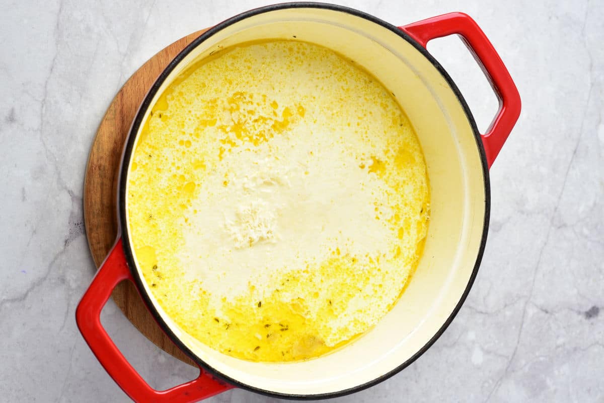 A large red and white pot filled with creamy yellow chicken gnocchi soup sits on a wooden trivet atop a light gray marble surface.