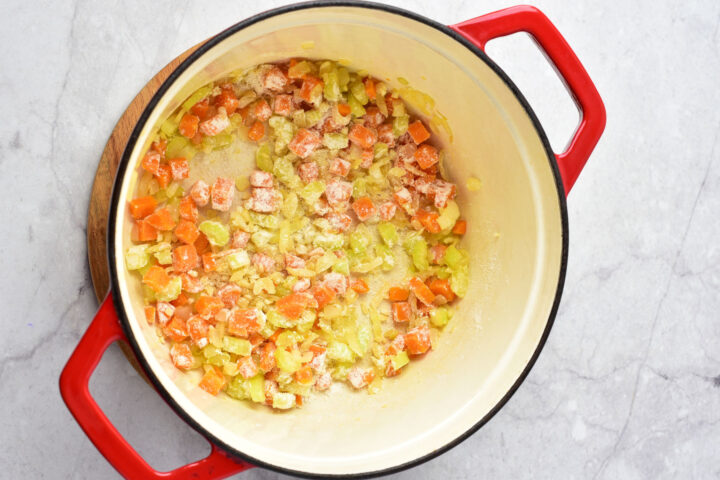 A red Dutch oven on a marble surface contains chopped onions, carrots, and celery being sautéed with flour sprinkled on top—the perfect base for a comforting chicken gnocchi soup.