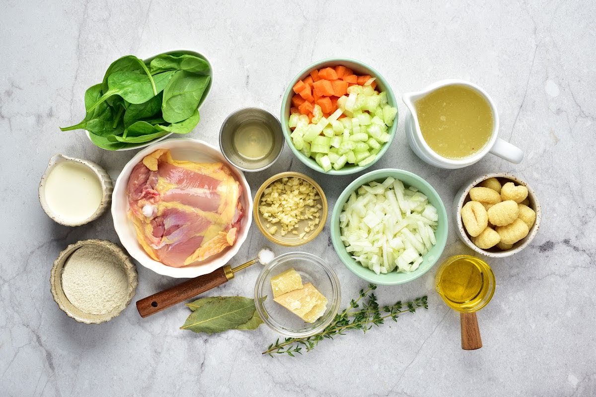 Various ingredients for a delicious chicken gnocchi soup are arranged on a white countertop, including raw chicken thighs, spinach, chopped carrots, celery, onions, gnocchi, broth, olive oil, garlic, grated cheese, heavy cream, seasonings, and herbs.