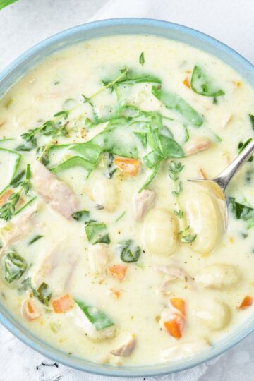 A bowl of creamy chicken gnocchi soup with spinach, carrots, and herbs, served in a blue bowl with a spoon, rests on a white napkin.