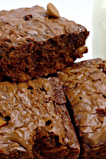 A close-up of rich, fudgy chocolate Elvis brownies containing bananas and bacon, stacked on a plate, with a glass bottle of milk in the background. The brownies have a shiny, crackly top.