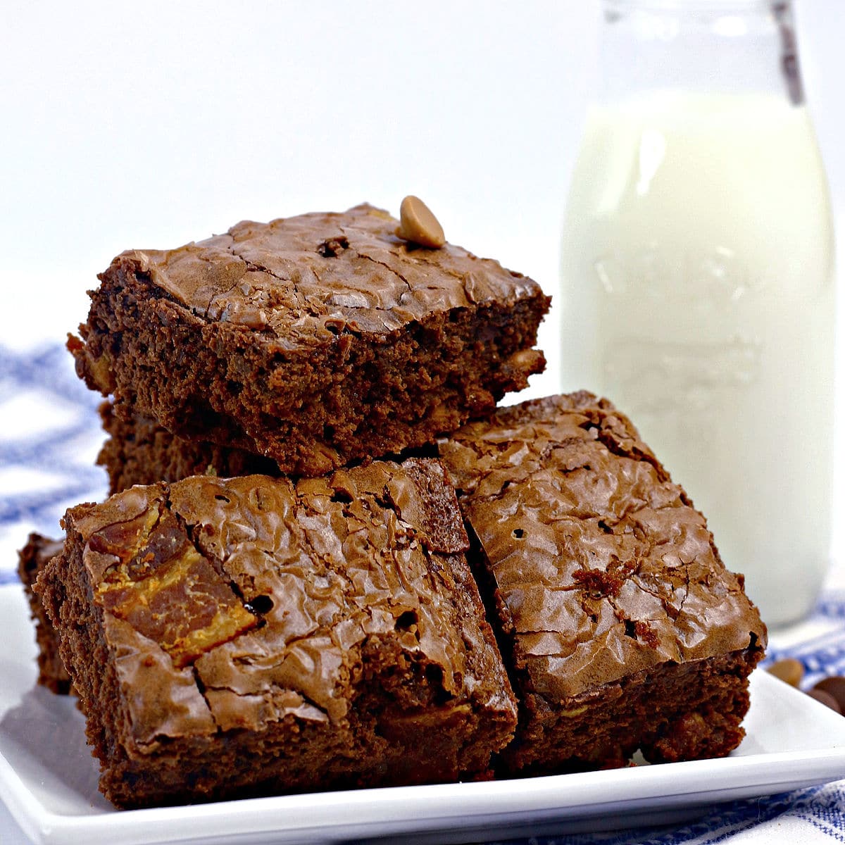 Three thick Elvis brownies stacked on a white plate, with a bottle of milk in the background. The brownies have a shiny, crackly top and rich, chocolatey texture.