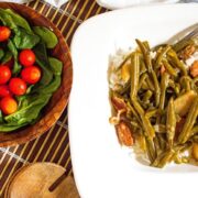 A square white plate with rice topped with green beans and sausage slices. Next to it is a wooden bowl containing fresh spinach leaves and cherry tomatoes, placed on a bamboo mat.