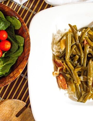 A square white plate with rice topped with green beans and sausage slices. Next to it is a wooden bowl containing fresh spinach leaves and cherry tomatoes, placed on a bamboo mat.