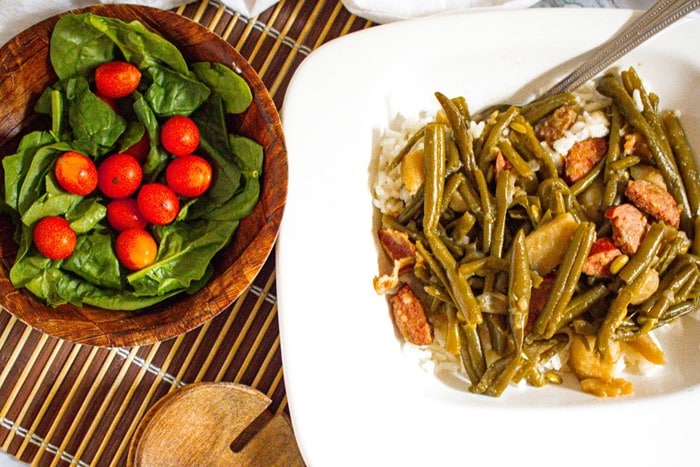 A square white plate with rice topped with green beans and sausage slices. Next to it is a wooden bowl containing fresh spinach leaves and cherry tomatoes, placed on a bamboo mat.
