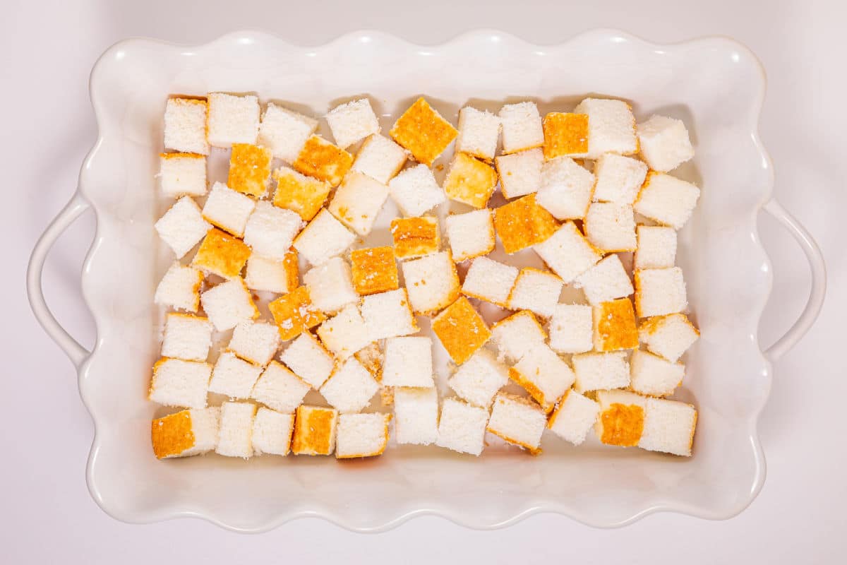 Cubed pieces of yellow cake with golden tops are evenly spread in a white ceramic baking dish with wavy edges and handles on each side.