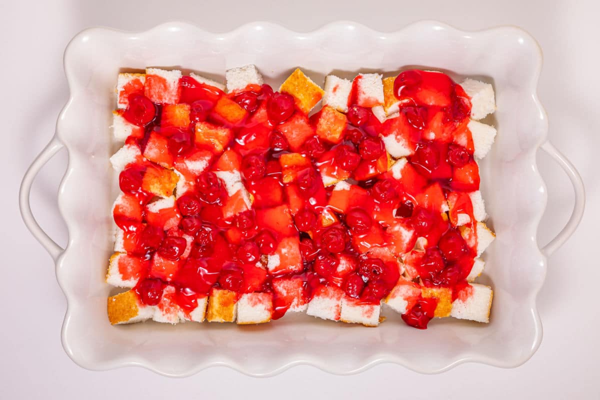 A white baking dish filled with cubes of yellow and white cake topped with bright red cherry pie filling. The dish has wavy edges and handles on both sides.