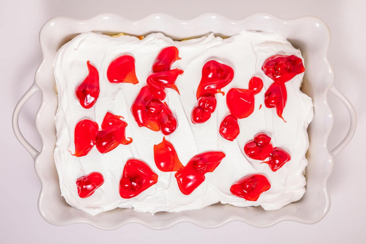 A rectangular white dish with a dessert topped with a thick layer of whipped cream and dollops of bright red cherry pie filling scattered across the surface.