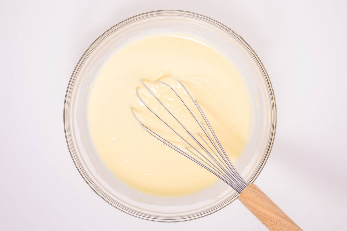 A glass bowl filled with pale yellow cake batter, with a metal whisk with a wooden handle resting inside, viewed from above on a white background.